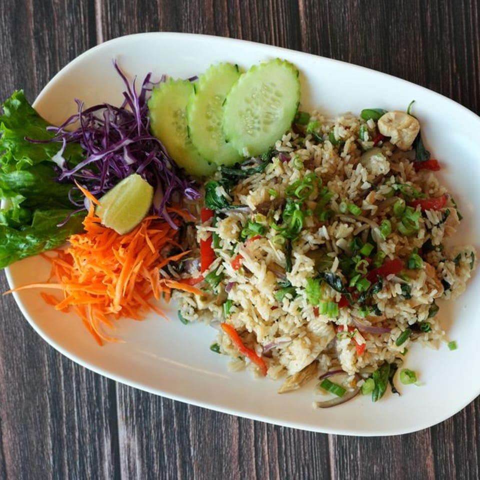 Thai fried rice with chicken, vegetables, lime, and cucumber slices on a white plate.